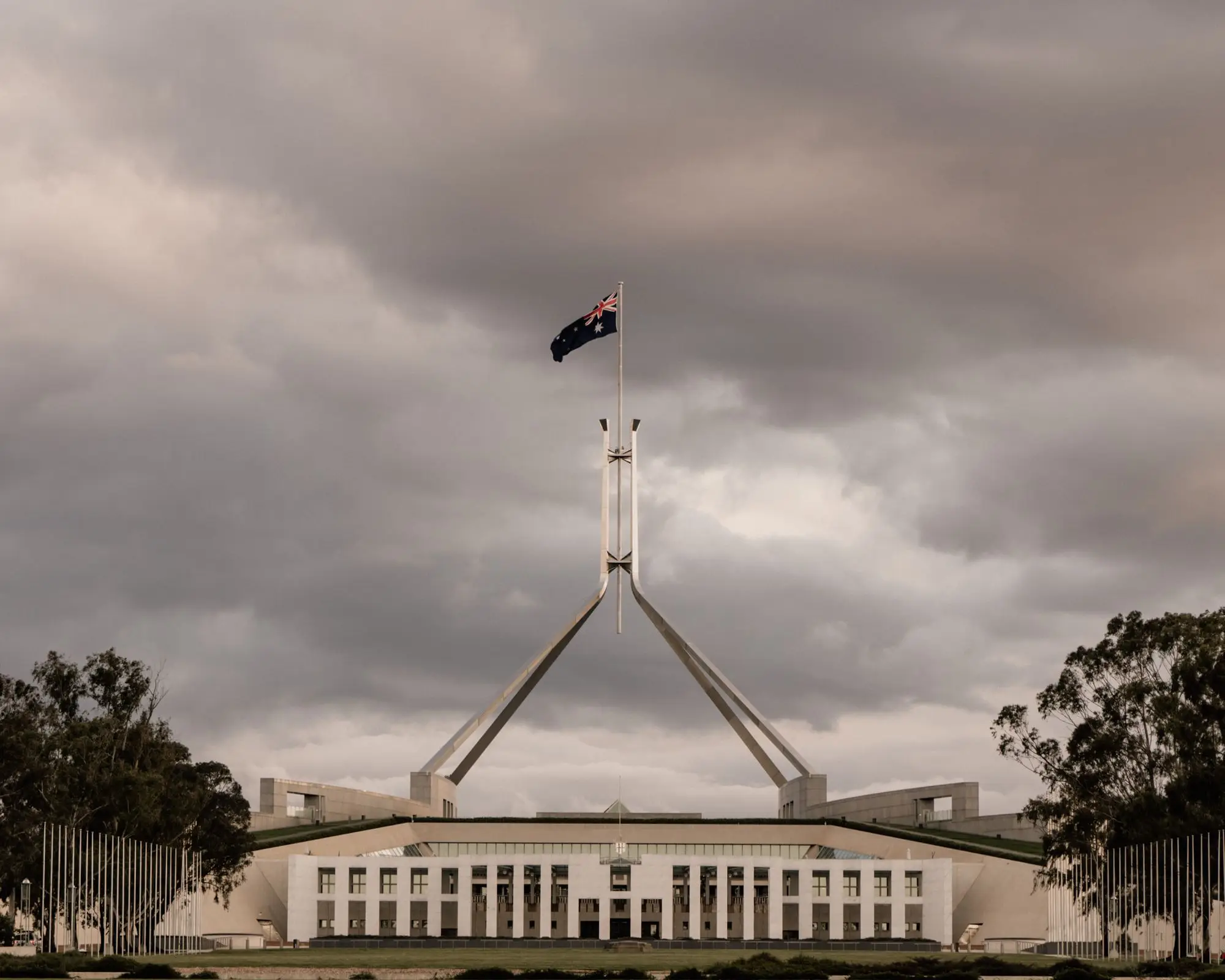 Parliament House in Canberra under heavy grey cloud, the Australian flag flying above the building
